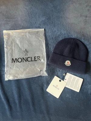 Mörkblå Moncler mössa  - Säljer en mörkblå ribbstickad mössa från Moncler med uppvikt kant och Moncler-logga framtill. Mössan är tillverkad i mjuk ull och kommer med originalpåse och tags kvar. Perfekt för kalla dagar och snygg till alla outfits.