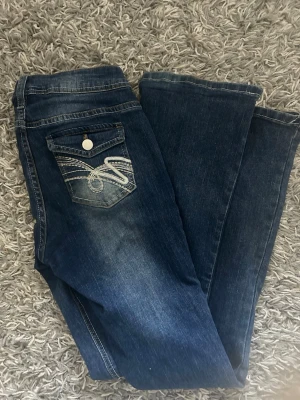 Mörkblå bootcut jeans med broderad ficka - Snygga mörkblå bootcut jeans med ljusa slitningar och broderad bakficka med vit söm. Klassisk femficksmodell och normal midja. Jeansen har en avslappnad passform och är tillverkade i denim med lite stretch för extra komfort. Det finns inga lappar därför vet jag inte var de är från men passar mig som är 164 och brukar ha 26/32 i andra jeans!