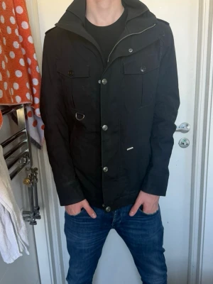 Svart jacka snoot  - Snygg svart vindjacka med både knappar och dragkedja framtill. Jackan har flera fickor med lock, hög krage och detaljer som metallring och axelklaffar. Passar perfekt till jeans och har en modern, clean look.