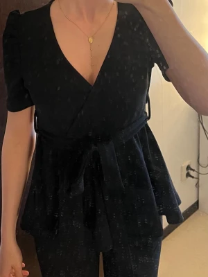 Svart omlottopp med knytband - Snygg svart omlottopp med v-ringning och korta puffärmar. Toppen har ett knytband i midjan som framhäver siluetten och ger en feminin look. Passar perfekt till både jeans och kjol för en trendig outfit.