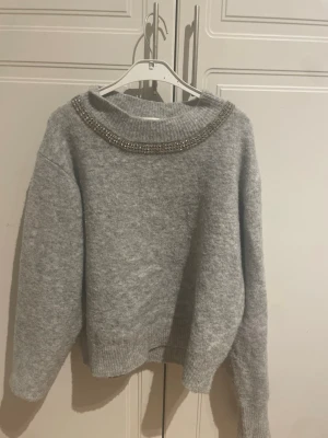 Grå stickad tröja med pärlor Kappahl - Mysig grå stickad tröja från Kappahl i storlek XS. Tröjan har en bred ribbad halsringning dekorerad med pärlor och stenar, vilket ger en extra touch. Långa ärmar och ribbade muddar vid ärmslut och nederkant. Perfekt för kalla dagar.