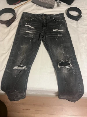 Supply demand jeans - Säljer ett par svarta jeans med slitningar och stora hål vid knäna. Jeansen har en tvättad look, raka ben och detaljer med mindre slitningar och färgstänk. Perfekta för en streetwear-stil och passar dig som gillar en edgy vibe.