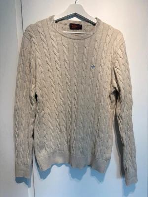 Beige tröja Gant - Kabelstickad beige tröja från Gant med rund halsringning och diskret blå logga broderad på bröstet. Riktigt snygg till en finare outfit. OBS: står att de är storlek XL men den är som M! Skriv vid fundering!