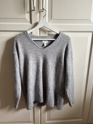 Grå stickad tröja från H&M - Mysig grå stickad tröja från H&M med v-ringning och långa ärmar. Tröjan har en avslappnad passform och ribbade muddar vid ärmslut och nederkant. Tröjan har två slitsar vid nederkant av tröjan på vardera sida (se bild 3)  Perfekt för lager på lager och enkel att matcha till olika outfits. 