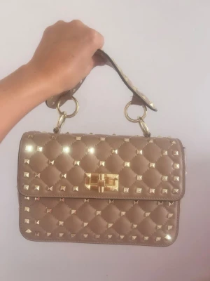 Beige valentino quiltad handväska med nitar - Stilren beige valentino inspererad väska  i quiltat mönster med guldfärgade nitar och matchande guldfärgat spänne. Väskan har ett kort handtag och rektangulär form. Perfekt accessoar för att lyfta din outfit.