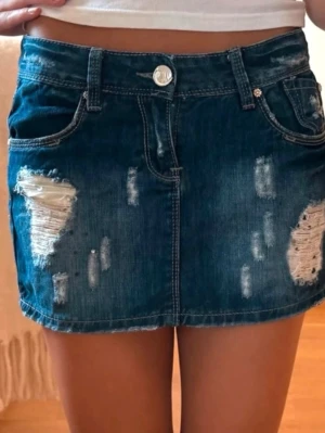Mörkblå jeanskjol med slitningar - Snygg mörkblå jeanskjol i kort modell med slitna detaljer och råa kanter. Kjolen har klassiska fickor fram och bak samt bälteshällor. Perfekt för dig som gillar en trendig och avslappnad stil.