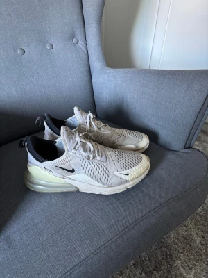Nike Air Max 270 beige sneakers - Säljer ett par Nike Air Max 270 sneakers i beige med svarta detaljer och vit sula. Skorna har en luftig mesh-överdel, synlig Air-enhet i hälen och snörning framtill. Perfekta för dig som gillar sportig stil och bekväma skor.