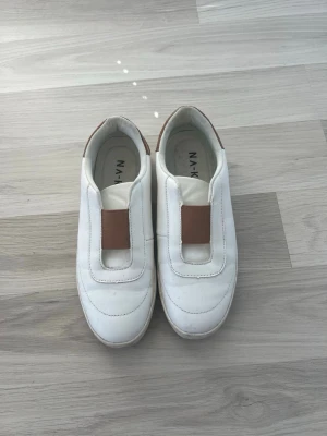 Vita slip-on sneakers med bruna detaljer - Snygga vita slip-on sneakers med bruna detaljer på häl och ovansida. Skorna har en stilren design utan snörning och är tillverkade i ett slätt material som ger en clean look. Perfekta för dig som gillar minimalistisk stil och vill ha något enkelt att dra på sig.