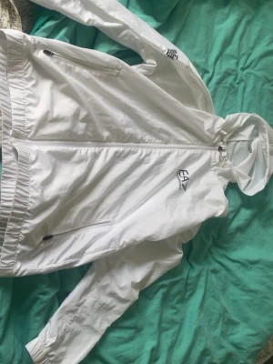 Vit vindjacka från Emporio Armani EA7 - Snygg vit vindjacka från Emporio Armani EA7 med huva och dragkedja framtill. Jackan har två sidofickor med dragkedja och elastiska muddar vid ärmslut och nederkant. Perfekt för dig som vill ha en stilren och sportig look.