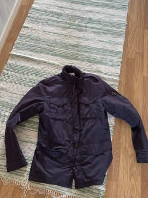 Mörkblå field jacket från J.Lindeberg - Snygg mörkblå fieldjacka från J.Lindeberg med hög krage och fyra framfickor med lock. Jackan har en patch med logga på ärmen och stängs med dragkedja och knappar. Tillverkad i ett lätt, vindtätt material som passar perfekt till blåsiga dagar.
