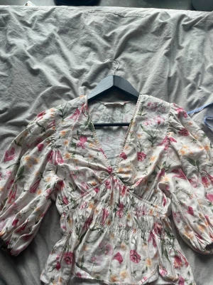 Blommig v-ringad blus med puffärm - Söt blus med v-ringning och puffiga långärmar. Blusen har ett blommigt mönster i rosa, grönt och gult på vit botten samt smock i midjan för en feminin siluett. Perfekt för dig som gillar romantisk och trendig stil.