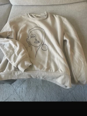 Beige sweatshirt med konstnärligt tryck - Snygg beige sweatshirt med ett stilrent, konstnärligt ansiktstryck i svart på framsidan. 