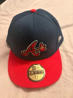 Blå och röd New Era 59FIFTY keps - Snygg 59FIFTY keps från New Era med Atlanta Braves-logga framtill. Kepsen är mörkblå med röd skärm och röd knapp på toppen. Broderad logga på sidan och klassisk flat brim. Perfekt för dig som gillar sportig streetstyle.