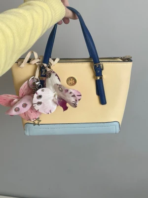 Pastellgul Handväska med Blå Detaljer - En superfin pastellgul väska med blå detaljer och söt fjärilsberlock. Perfekt till vår‑ och sommaroutfits, matchar både denim och ljusa färger. Rymlig nog för mobil, plånbok och småsaker. Använd få gånger och i jättefint skick. Pris kan diskuteras vid snabb affär.
