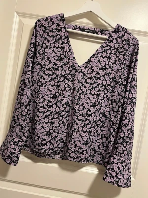 Lila blommig blus med v-ringning - Snygg blus med lila blommönster på svart botten och v-ringad hals. Blusen har långa ärmar och ett avslappnat fall. Perfekt för dig som gillar en feminin och trendig stil.