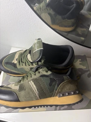 Valentino Garavani camo sneakers - Säljer ett par snygga sneakers från Valentino Garavani i camomönster. Skorna har gröna, svarta och beige toner, detaljer i mocka och mesh samt nitar på hälen. Sulan är i gummi med beige färg och svart kant. Perfekta för dig som vill sticka ut med stil.