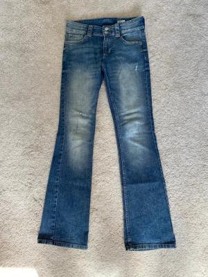 Blå bootcut jeans - Säljer ett par blå bootcut jeans med snygga slitningar och ljusare partier på låren och knäna. Jeansen har klassisk femficksmodell, knapp- och dragkedjegylf samt fickor med lock bak. 