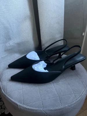 Svarta spetsiga slingback-pumps - Säljer ett par stilrena svarta pumps med spetsig tå och låg klack. Skorna har öppen häl med smal rem runt ankeln och är tillverkade i ett slätt material som ger en elegant look. Perfekta för dig som gillar klassisk och trendig stil.