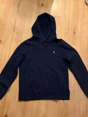 Mörkblå hoodie från Polo Ralph Lauren - Snygg mörkblå hoodie från Polo Ralph Lauren med klassisk huva och känguruficka. Tröjan har det ikoniska broderade logotypen på bröstet och är tillverkad i mjukt bomullsmaterial. Perfekt för en avslappnad och stilren look. Den är utvetad som man se på bilderna storlek 18/20. L barn men passar xs/s i vuxen 