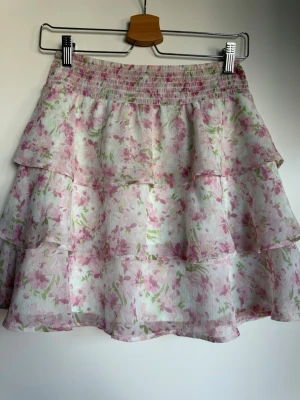 Vit volangkjol med blommigt mönster - Söt kjol i vitt med rosa och gröna blommor. Kjolen har flera volanger och resår i midjan för en bekväm passform. Tunt, luftigt material som ger en lätt och somrig känsla. 