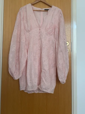 Ljusrosa  klänning från  Gina Tricot - Söt ljusrosa klänning från Gina Tricot med broderade blommor och hålmönster. Klänningen har v-ringning, lång ärm och volangkant vid halsen. Perfekt för sommarens alla tillfällen.