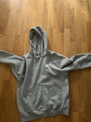 Grå hoodie från Nike med huva - Säljer en klassisk grå hoodie från Nike med vit logga på bröstet. Hoodien har stor känguruficka framtill och en rejäl huva med dragsko. Perfekt för chill dagar eller när du vill ha en enkel och snygg look. Mjuk bomullsblandning och ribbade muddar.