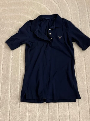 Mörkblå pikétopp med krage - Klassisk mörkblå pikétopp med krage och knappar framtill. Toppen har korta ärmar och liten logga på bröstet. Perfekt till jeans eller kjol för en avslappnad look.