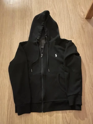 Svart zip up från Polo Ralph Lauren - Säljer en svart hoodie från Polo Ralph Lauren med dragkedja, huva med dragsko och fickor framtill. Klassisk vit logga broderad på bröstet. Perfekt för en chill och stilren look. Tillverkad i mjukt material för extra komfort.