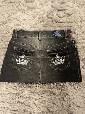 Svart jeanskjol med kronbrodyr - Cool svart jeanskjol med slitna kanter och två bakfickor med vit kronbrodyr. Kjolen har klassisk femficksdesign, bälteshällor och knappstängning framtill. Perfekt för en trendig och avslappnad look.