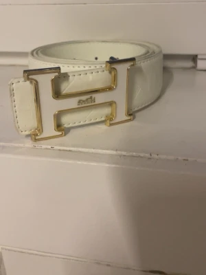 Vitt skärp med guld H-spänne - Snyggt vitt bälte med ett stort H-format spänne i guld och vitt. Bältet är 110 cm långt och har en stilren look som passar till många outfits. Perfekt accessoar för dig som vill sticka ut lite extra.