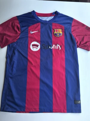 Barcelona fotbollströja  - Snygg Barcelona matchtröja från Nike i blått och rött med klubbmärke och Spotify-logga på bröstet. Tillverkad i lätt och ventilerande material, perfekt för fotboll eller träning. Klassiska vertikala ränder och korta ärmar.
