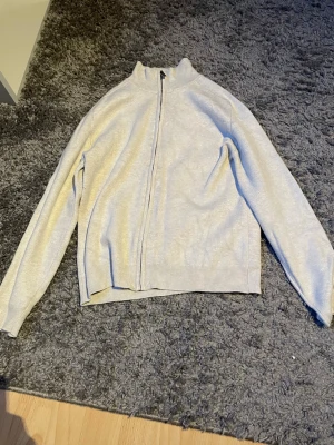 Ljusgrå ziptröja från Lindex - Ljusgrå ziptröja från Lindex med hög krage och hel dragkedja framtill. Tröjan har ribbade muddar vid ärmslut och nederkant, samt en enkel och stilren design som passar till det mesta. Perfekt för lager på lager och enkel att matcha.
