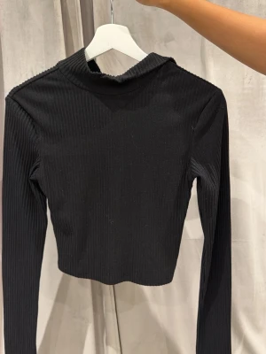 Svart ribbad croppad polotopp - Snygg svart croppad polotopp med ribbad struktur och långa ärmar. Toppen har en hög krage med knappar som kan öppnas för en cool detalj. Perfekt att styla med jeans eller kjol för en trendig look.
