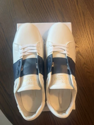 Valentino Garavani - Snygga vita sneakers från Valentino med bred svart läderdetalj över mitten och vita skosnören. Skorna har en klassisk låg profil och diskret logga på plösen. Tillverkade i slätt läder för en clean och exklusiv look.