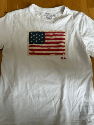 Vit t-shirt Polo Ralph Lauren USA-flagga - Vit t-shirt från Polo Ralph Lauren med tryck av amerikanska flaggan på bröstet och diskret RL-signatur. Klassisk passform och rund halsringning. Perfekt för dig som gillar stilrena plagg med en twist.