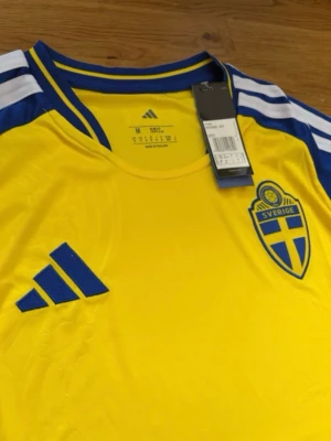 Svensk landslagströja Adidas gul/blå - Gul svensk landslagströja från Adidas med blå detaljer och vita ränder på axlarna. Tröjan har broderat Sverige-emblem på bröstet och Adidas-logga. Tillverkad i lätt och funktionellt material, perfekt för fotboll eller träning.