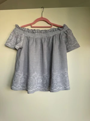 Randig offshoulder topp med broderi - Säljer en söt offshoulder topp med smala blå och vita ränder. Toppen har korta ärmar, broderade detaljer längst ner och en luftig passform. Perfekt för varma dagar och ger en chill sommarkänsla.