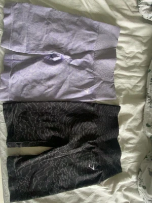 Två par seamless shorts i lila och svart - Två par seamless shorts, ett par i ljuslila och ett par i svart med diskret mönster. Båda har hög midja och stretchigt material som passar perfekt till träning. Liten logga framtill på båda paren. De har storleken S men kan sitta åt lite om midjan. Hade även passat någon med storleken XS. Beror på hur man vill att de ska sitta 