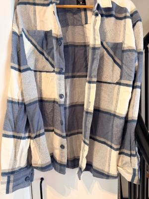 Blåvit rutig skjorta med knappar - Säljer en blåvit rutig skjorta med långa ärmar och två bröstfickor. Skjortan har klassisk krage och knäppning framtill. Perfekt för dig som gillar en avslappnad och trendig stil.