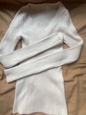 Vit ribbad långärmad tröja - Säljer en vit, ribbstickad tröja med lång ärm och djup v-ringning. Tröjan är figurnära och har en stretchig passform som framhäver siluetten. Perfekt att styla med jeans eller kjol för en clean look.