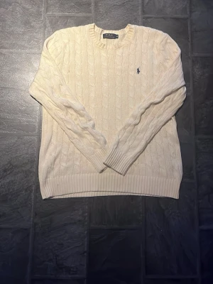 Biege  kabelstickad tröja från Polo Ralph Lauren - Kabelstickad vit tröja från Polo Ralph Lauren med klassisk rund halsringning och ribbade muddar. Tröjan har den ikoniska mörkblå logotypen broderad på bröstet och är tillverkad i mjukt material. Perfekt för en clean och stilren look.