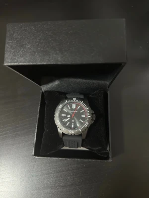 Svart klocka från Bruno Banani - Snygg svart klocka från Bruno Banani med sportig design. Klockan har svart urtavla, vita markeringar och detaljer samt en röd sekundvisare. Armbandet är i svart syntetmaterial och boetten är robust med tydliga siffror på ringen.