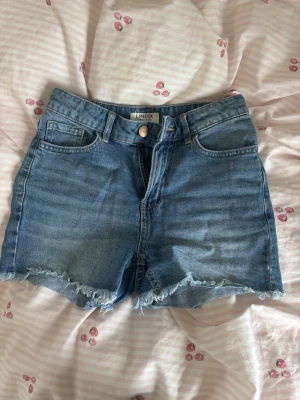 Blå jeansshorts från Lindex - Snygga blå jeansshorts från Lindex med råa kanter och klassisk femficksmodell. Shortsen har hög midja och stängs med knapp och dragkedja. Perfekta för varma dagar och enkla att matcha med olika toppar.