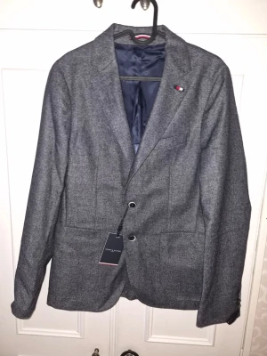 Blå kavaj med två knappar från Tommy Hilfiger  - Stilren blå kavaj med två knappar framtill och klassisk krage. Kavajen har en liten broderad detalj på bröstet och är helfodrad med mörkblått foder. Passar perfekt till skjorta och jeans för en snygg look. Nypris: 3500kr