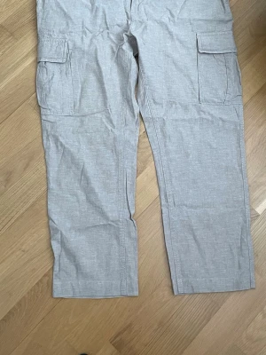 Beige cargobyxor med fickor, 36/30 - Snygga beigea  cargobyxor med raka ben och stora sidofickor. Byxorna har knappdetaljer bak och är tillverkade i ett lätt bomullsliknande material. Perfekta för en avslappnad och trendig stil. De har förkortats i längden så fråga  vid intresse .
