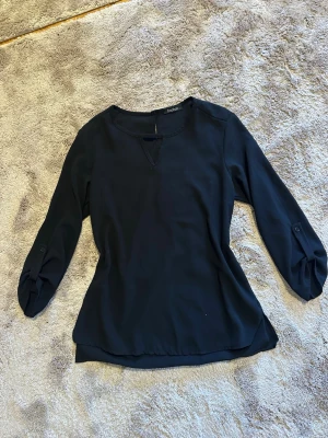 Svart blus med knappdetaljer - Stilren svart blus med rund halsringning och liten V-detalj framtill. Blusen har långa ärmar med knappdetaljer vid ärmsluten och är tillverkad i ett lätt,  tyg som ger en snygg passform. Perfekt att matcha med jeans eller kjol.
