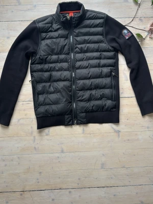 Svart dunväst från Parajumpers - Snygg svart dunväst från Parajumpers med quiltad front och stickade ärmar. Västen har dragkedja framtill, två sidofickor med dragkedja jackan är fake och har ingen gul flärp vid nacken 