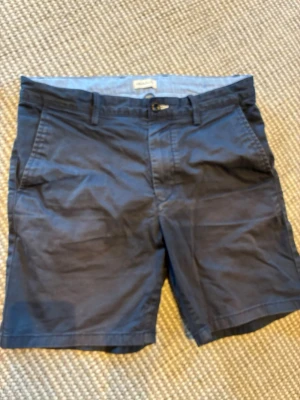 Mörkblå shorts från GANT - Snygga mörkblå shorts från GANT i klassisk modell med fickor på sidorna och bälteshällor. Tillverkade i mjuk bomull som känns skön mot huden. Perfekta för varma dagar och enkel att matcha med olika stilar. Se foton för mindre fläck.