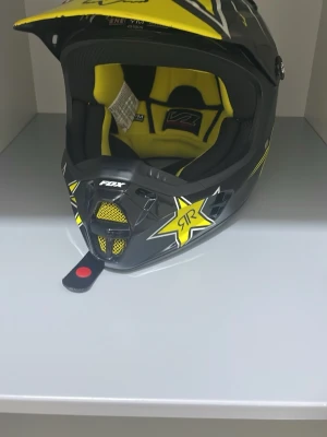 Svart och gul crosshjälm FOX - Säljer en svart och gul crosshjälm från FOX med Rockstar Energy-stjärnor på sidorna. Hjälmen har ett coolt mönster, ventilationsnät framtill och vadderad insida i gult och svart. Perfekt för motocross eller enduro. Den är osynlig sista gången man sålde den ny var det i 2013
