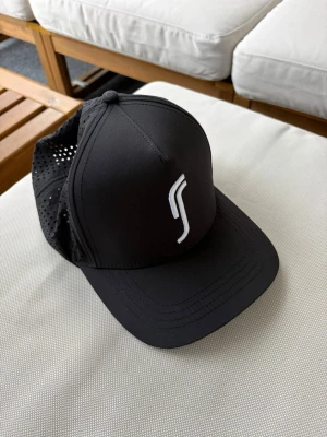 Svart keps med vit logga - Snygg svart keps med böjd skärm och vit broderad logga framtill. Baksidan är i mesh för extra ventilation och kepsen har justerbar storlek. Perfekt för en sportig och avslappnad stil.